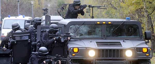 A swat team in black tactical gear hunkers behind a humvee, ready to fire on a target