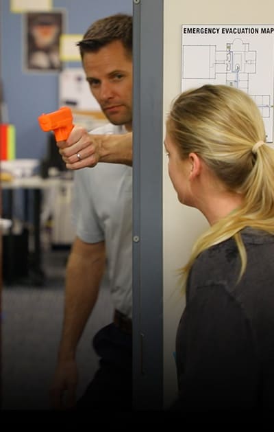 A man with a fake orange gun is hiding behind a door frame, simulating active shooter training